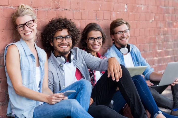 Portrait of friends sitting against wall using mobile phone, digital tablet and laptop