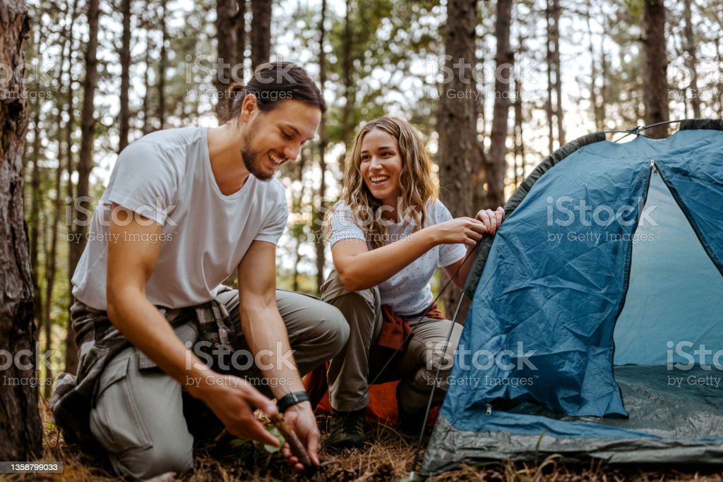 Beautiful smiling women with handsome boyfriend setting up tent in forest while talking